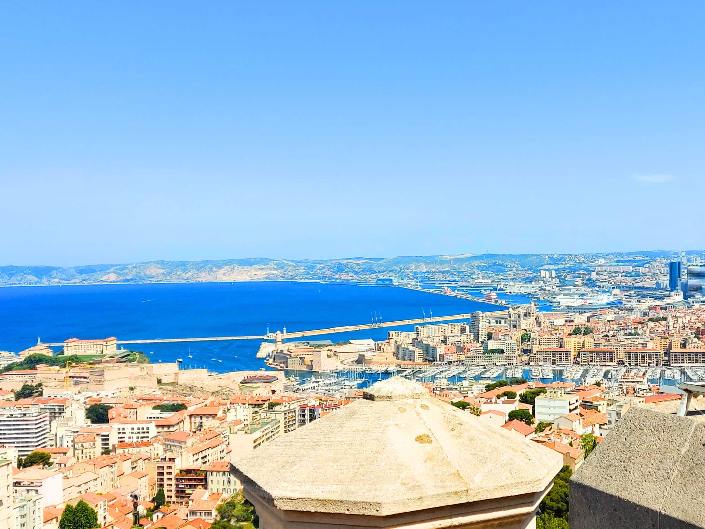 Panoramic view of a coastal city with a bustling harbor and deep blue waters, surrounded by distant mountains under a clear blue sky.