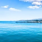 A calm, expansive blue sea with a distant shoreline and mountains under a bright, clear sky, capturing the serene beauty of a Mediterranean coastal landscape.