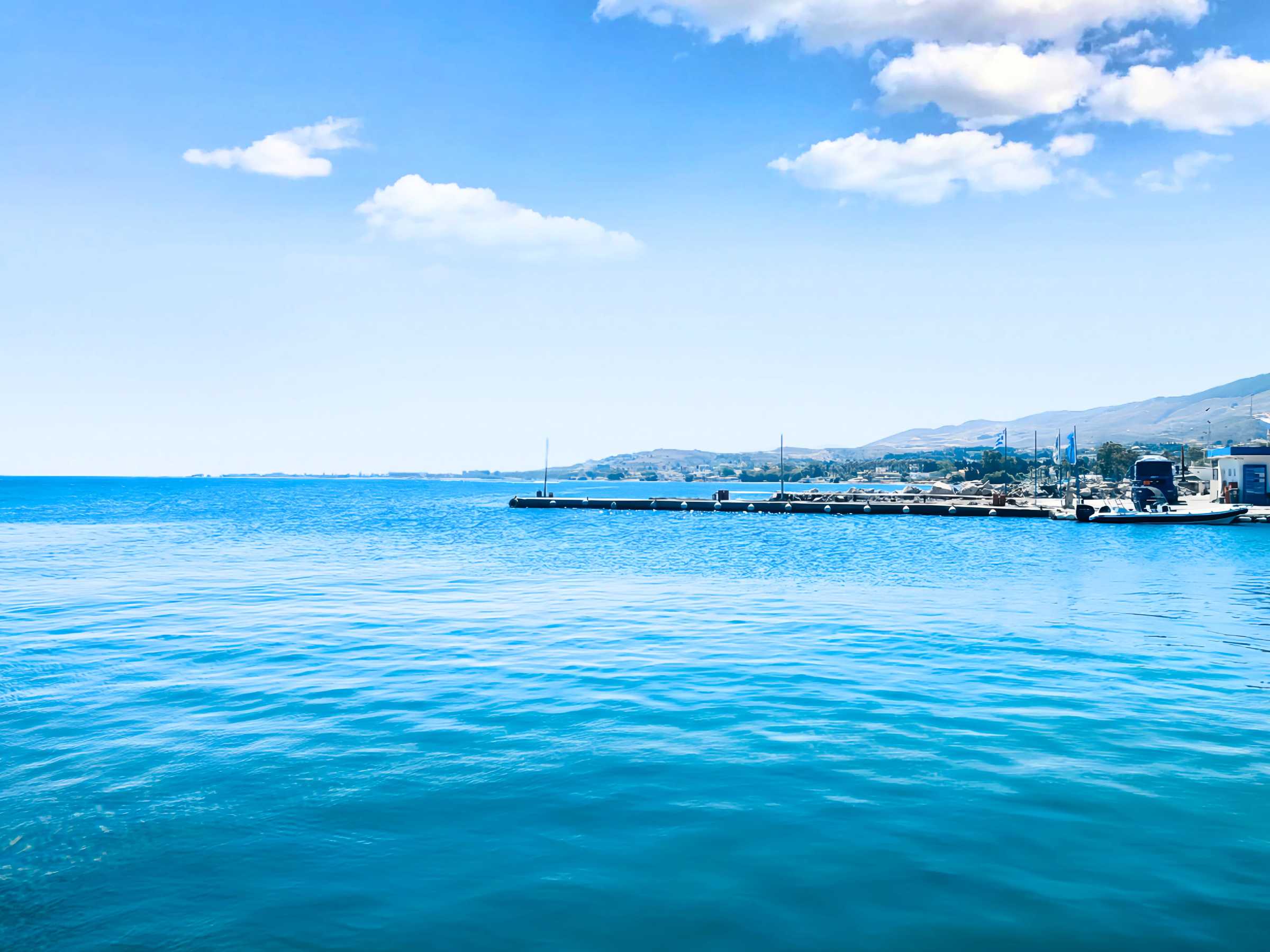 A calm, expansive blue sea with a distant shoreline and mountains under a bright, clear sky, capturing the serene beauty of a Mediterranean coastal landscape.