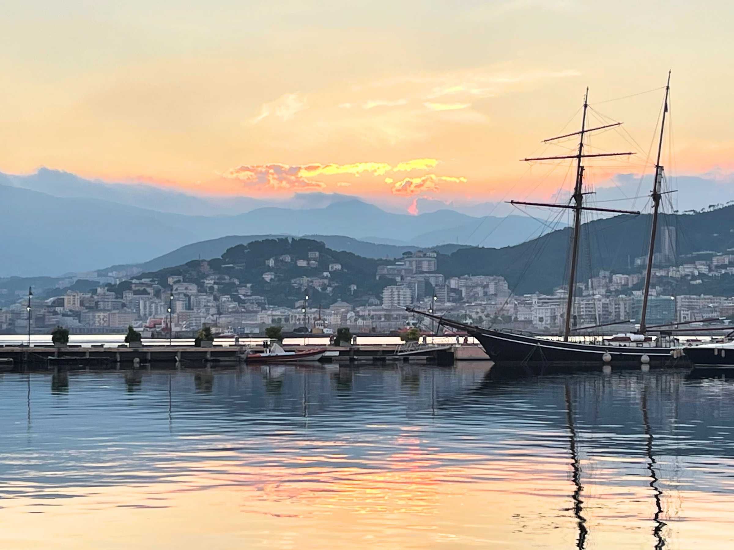 A sailboat docked in a calm harbor at sunset, with the orange and pink hues of the sky reflecting on the water, and a coastal city and mountains in the background, capturing the essence of sailing the Mediterranean.