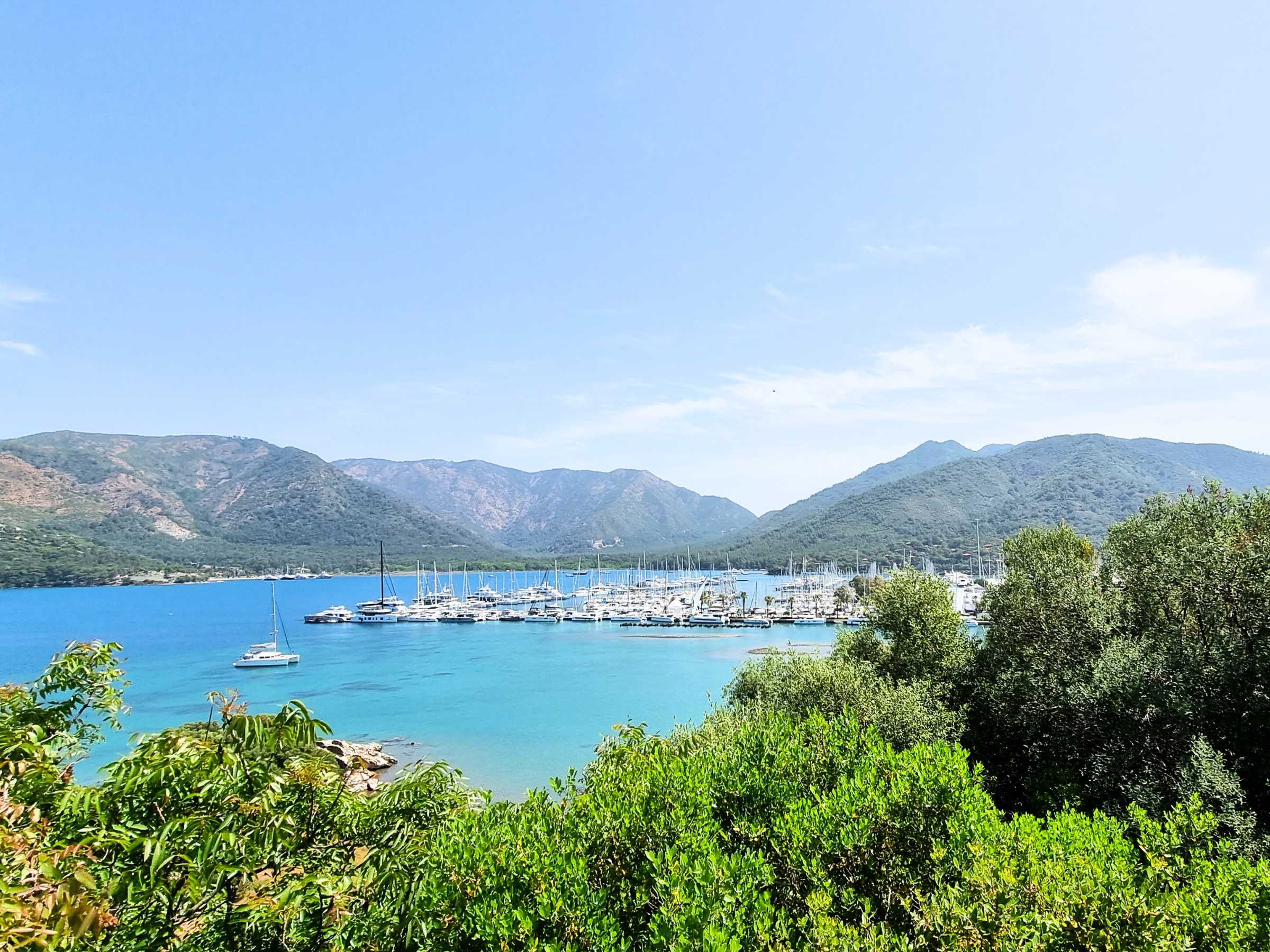 A picturesque marina nestled in a turquoise bay surrounded by lush green hills and distant mountains under a clear blue sky, capturing the essence of sailing the Mediterranean.